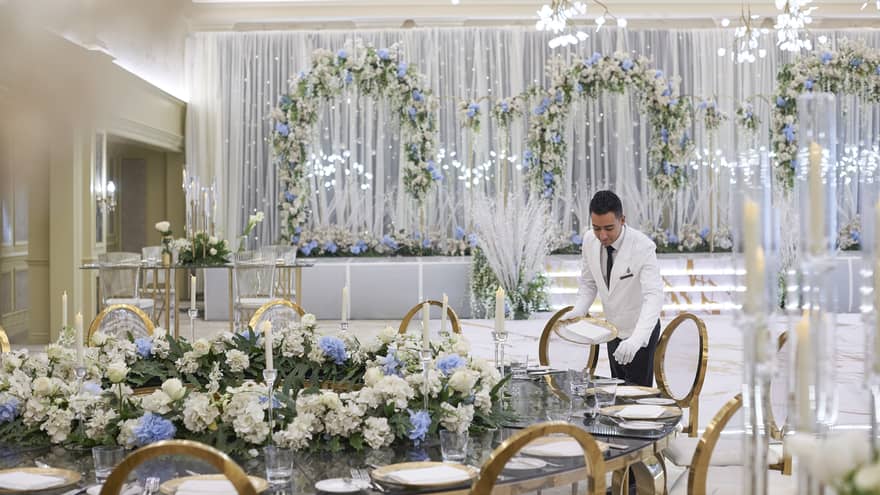 Formally dressed employee puts final touches on elegant wedding setup amidst grand floral decor