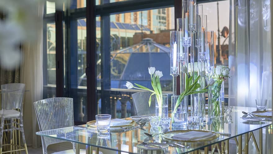 Elegantly set glass-top dining table, clear chairs, against black picture window