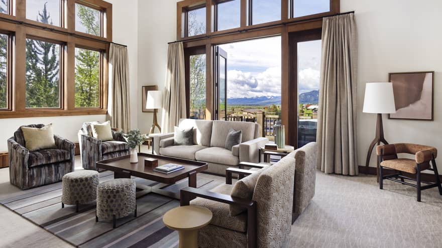 Luxury resort living room with high ceilings, large windows, sofa, armchairs, ottomans, table, floor lamps and doors opening to a mountain-view balcony