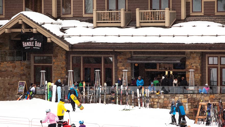 People with skis, snowboards walk up to Handle Bar entrance, skis lean against stone wall
