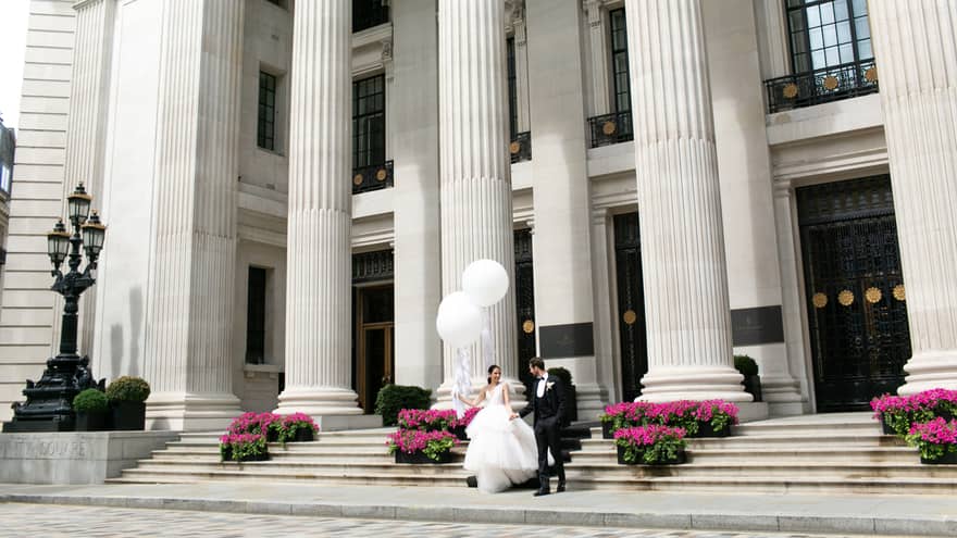 A bride and groom holding umbrellas, gracefully descending large stone steps outside historic building with grand white pillars