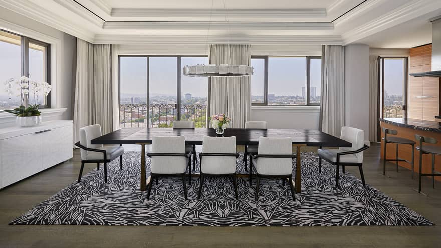 Large dark wood dining table with 10 white chairs, abstract rug, crown molding, hanging light and wall of windows with city view