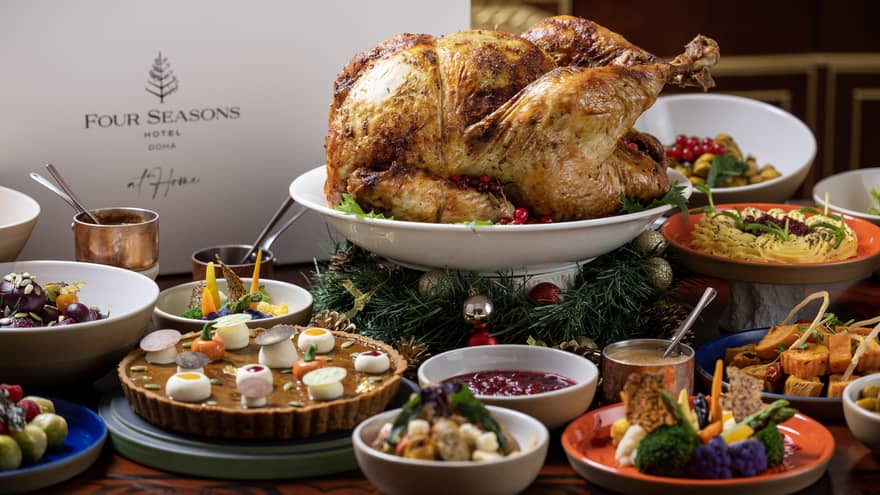 A whole roasted turkey on a white round platter sits elevated in the middle of a table filled with various bowls and plates of side dishes and sauces