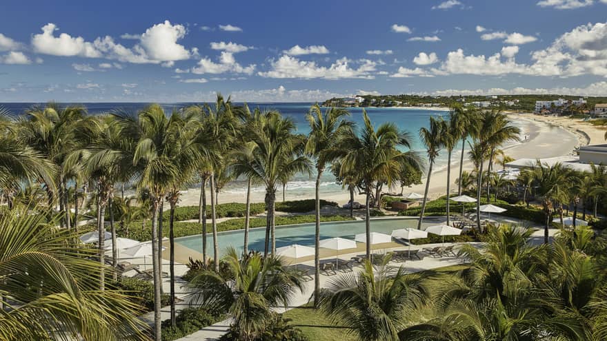 Aerial view of Bamboo Pool surrounded by tall palms, white patio chairs and umbrellas, steps from sand beach and ocean