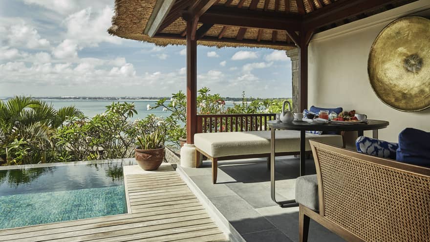Close-up of Deluxe Villa patio corner with swimming pool, plant, white bench under thatched roof, ocean view