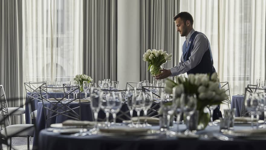 A four seasons male staff placing a white floral arrangement onto a circular table, draped in a dark gray table-cloth