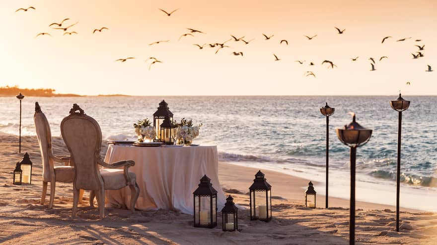 Romantic beachside dining setup with vintage chairs, a table decorated with flowers and lanterns and birds flying overhead