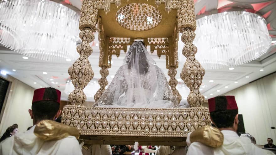 Wedding ceremony, back view of bride with white veil seated on beaded roofed platform carried by men