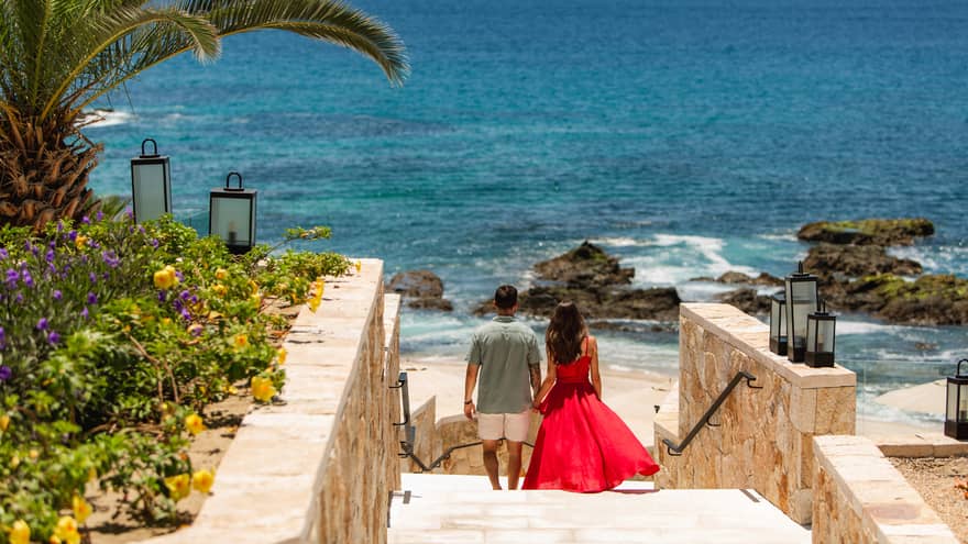 A man and woman walk down stairs holding hands, toward a tropical beach