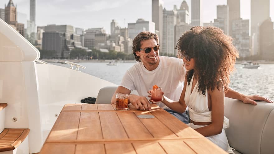 A man and woman sitting on a boat drinking cocktails with a city in the distance.