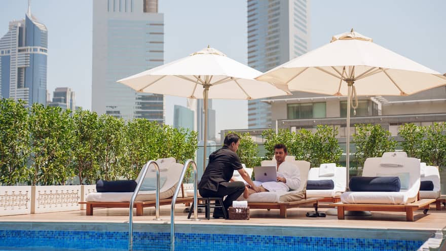 Guest sits on a poolside lounge chair looking at his laptop while a spa attendant rubs his feet