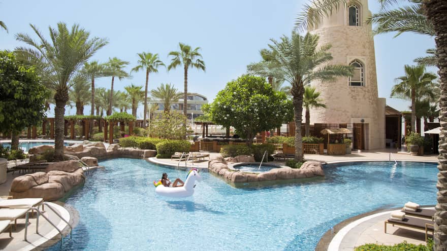 A guest lounges in a unicorn-shaped inner tube floating in the outdoor pool, flanked by palm trees and greenery