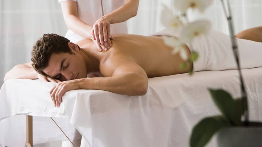 Man lays on white table with eyes closed as masseuse massages his bare shoulders