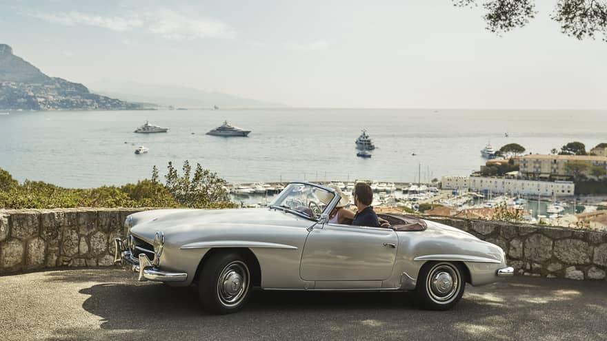 Couple sits in silver vintage convertible car parked on road by stone wall, looking over sea 