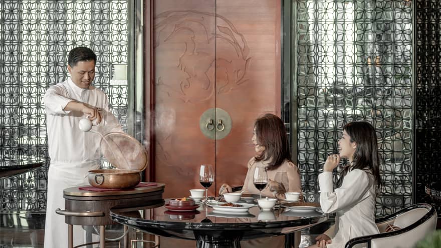Two female diners seated at table watch as chef pours liquid into large copper pot 