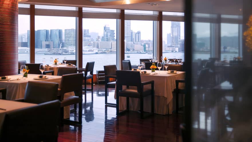 A sophisticated restaurant with neatly set tables and chairs. Large windows offer panoramic views of the city skyline and the waterway. 