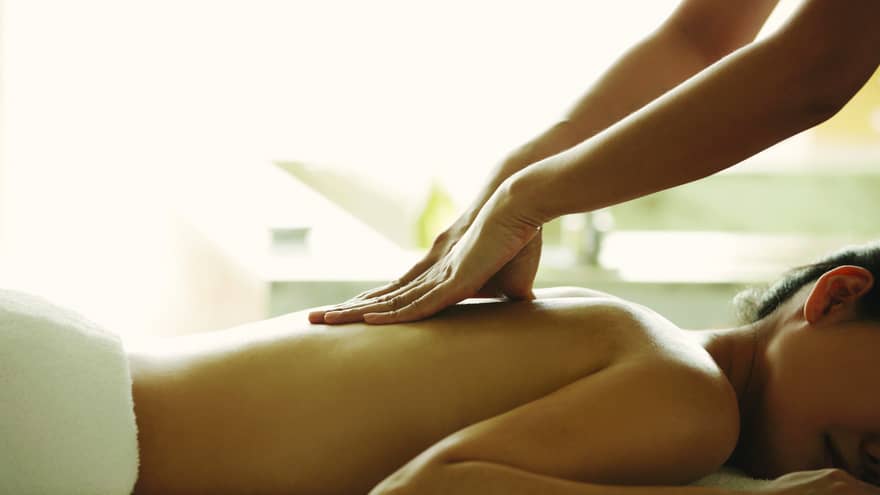 Masseuse reaches over, massages woman's bare back as she lies on spa table