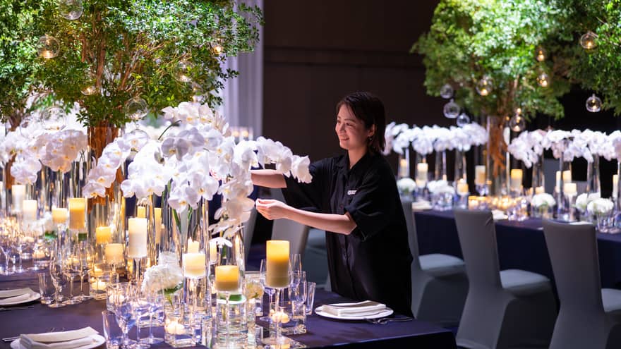 A wedding staff member preparing a table with elegant glassware and candlelights, arranging the white floral centrepiece to create a fresh and inviting setting.