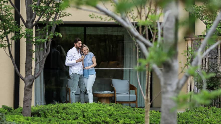 A couple stands in front of a sleek glass door on their hotel room balcony, holding champagne glasses, gazing at greenery outside.