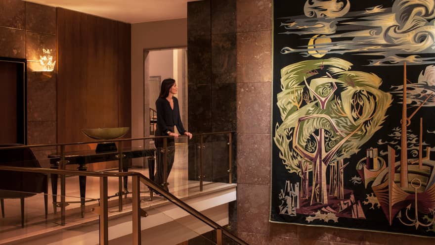 Woman dressed in black stands at glass railing in hotel lobby, looks at large colourful tapestry