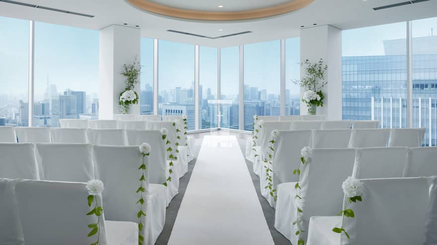 An indoor wedding venue with rows of white chairs and flowers, with a view of the city skyline from window.