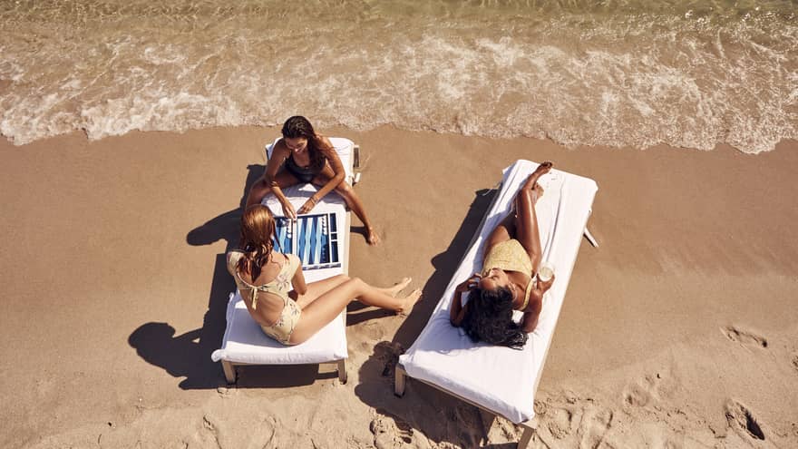 People relaxing on lounge chairs at a sandy beach, with two of them playing a backgammon game