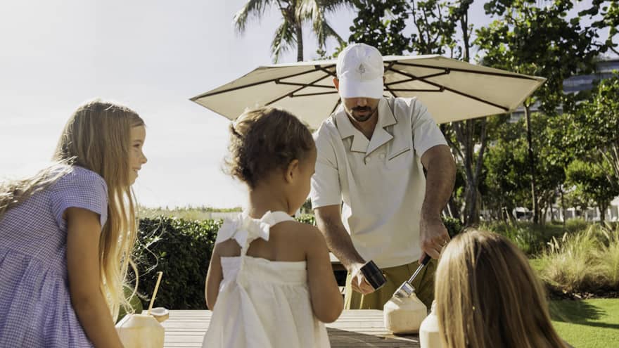 Man cracks a coconut as kids watch on beachfront lawn