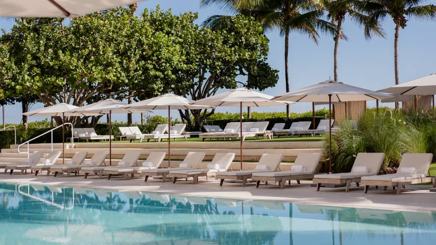 Luxury hotel outdoor pool area with rows of white lounge chairs, umbrellas and neatly landscaped greenery in the background