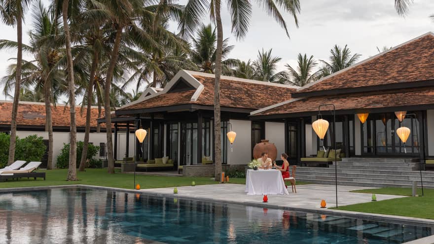 A couple enjoying a romantic dinner by a pool at a luxury villa, surrounded by palm trees and illuminated by hanging lanterns.