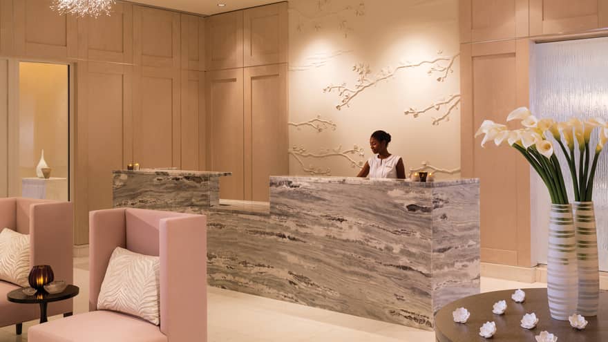A well lit lounge of a hotel with light pink chairs for people to rest and a worker behind a grey and white desk area.