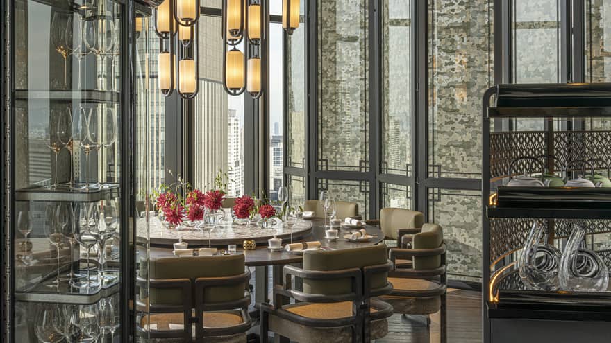 A mod chandelier hangs over red chrysanthemums on a round dinner table set beside tall windows and a vitrine of wine glasses.