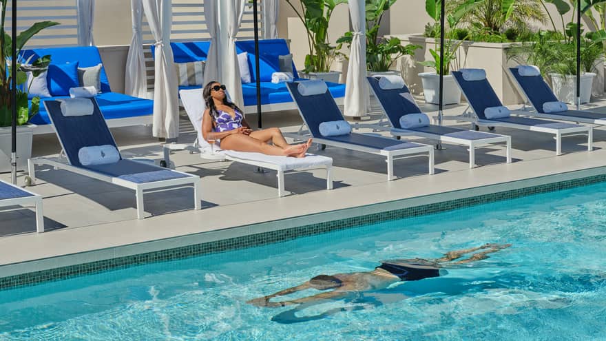 One guest laying on a lounge chair while another guest swims in the pool.