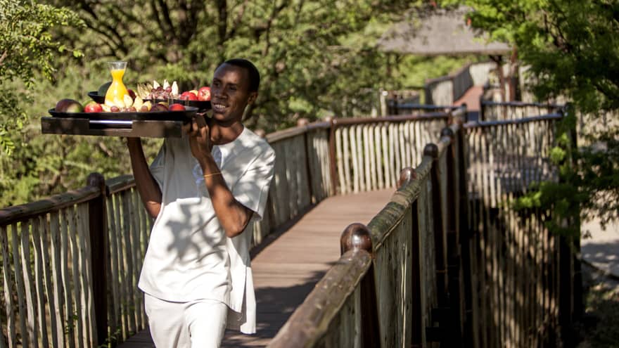 Hotel staff carries tray with fresh fruit, juice along wood bridge