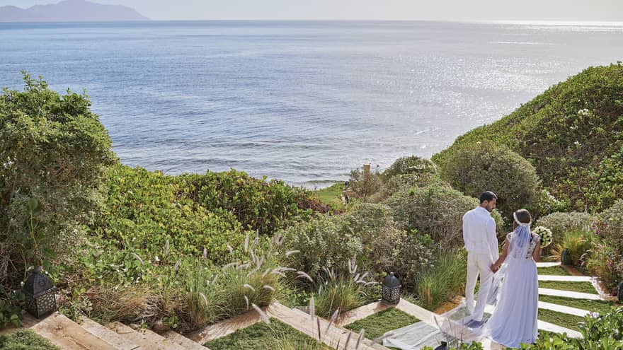 Couple walk down lawn steps through trees towards sea 