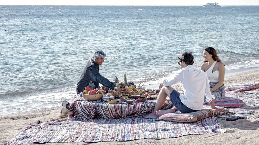 Three people sit on a beach around a low table set with a luxurious array of fruit, bread, and clay dishes full of food.
