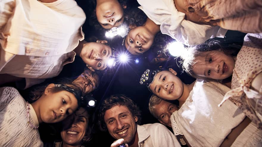 A group of children and adults, some wearing headlamps, stand in a circle and smile downwards into the camera. 