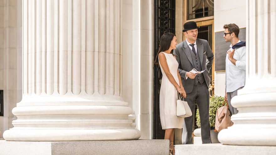 A couple converses with a Concierge on sunny steps beneath a large white pillar outside Four Seasons Hotel London at Ten Trinity Square.