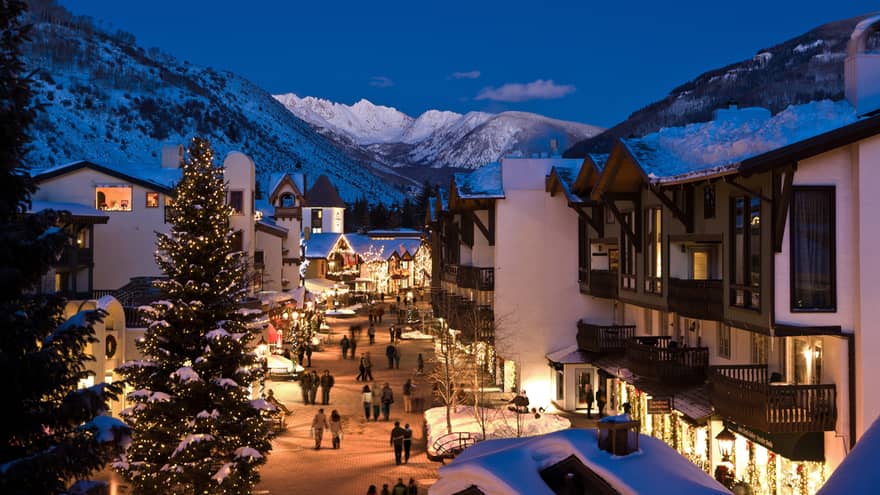 A snow covered hotel property surrounded by mountains and trees with lights coming out of buildings and street lamps at night.