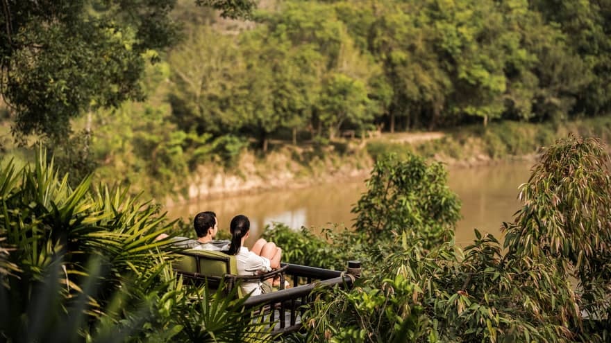 Couple sits on wood bench surrounded by tropical plants, overlooking river