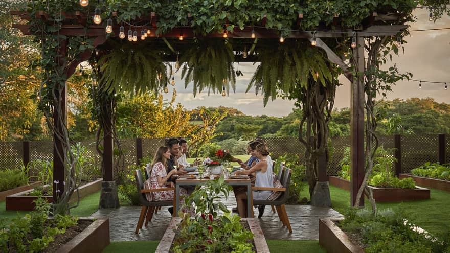 A group of people sit at an outdoor table set beneath a pergola draped in greenery and string lights in the middle of the resort garden