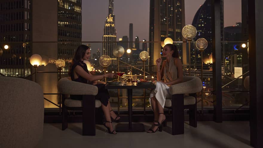 Two women sit at a cocktail table on an outdoor rooftop patio overlooking the Dubai skyline at night