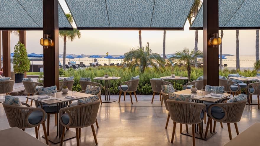 Covered restaurant terrace with grey and blue tables decor leads out to an open-air deck with greenery and the beach in the distance