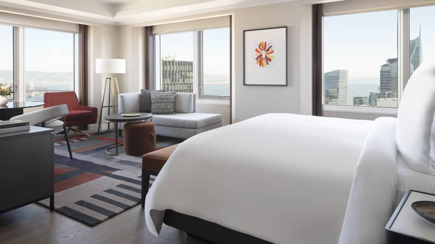 A hotel room with a large bed, large windows on each wall, a sofa, desk area, and lamp.