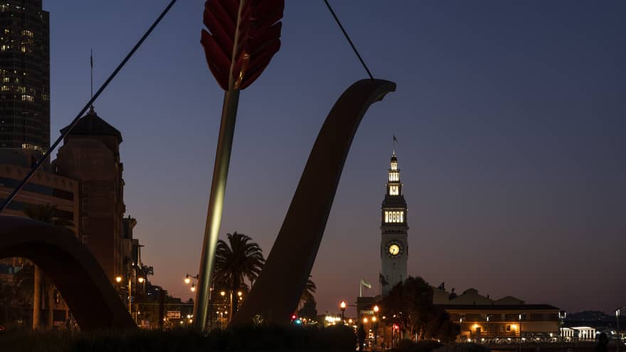 A nighttime image of a city with a clock tower, a large sculpture of a bow and arrow and building.