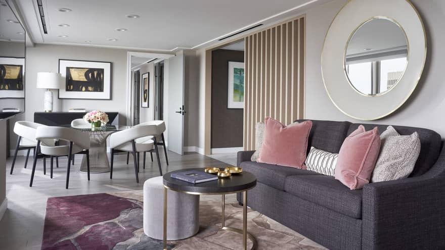 A hotel room with dark grey sofa, pink and grey pillows, coffee table, mirror and small table with chairs.