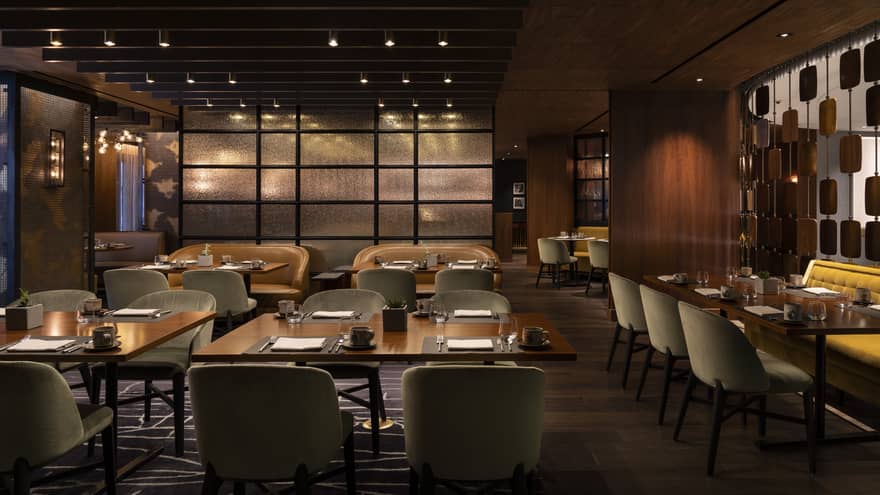 The interior of a dimly lit restaurant with square tables, grey chairs and wood walls.