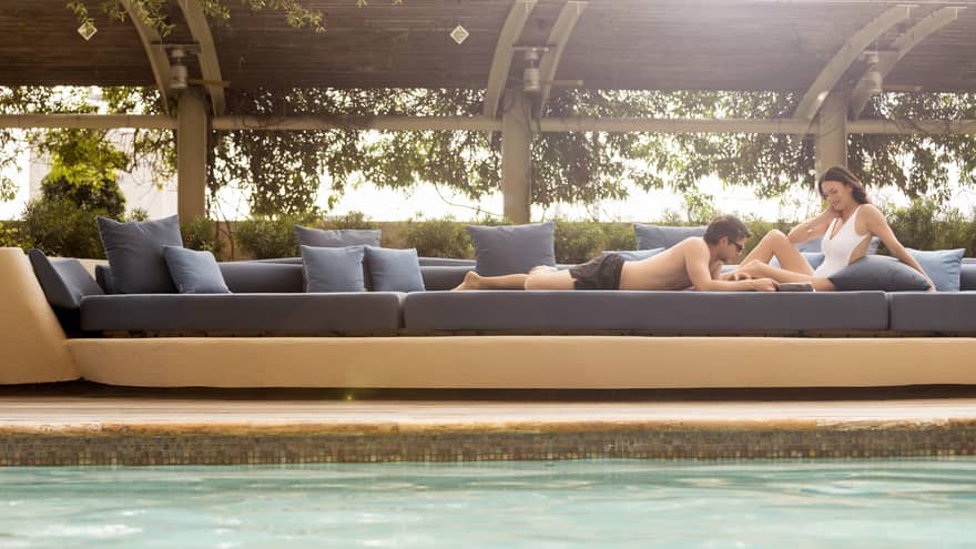 Man in swim trunks and woman in white swimsuit sunbathe on large blue patio banquette 