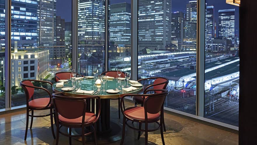 Luxury boutique hotel dining setup with a round table elegantly set for guests, offering a panoramic city view at night through tall glass windows