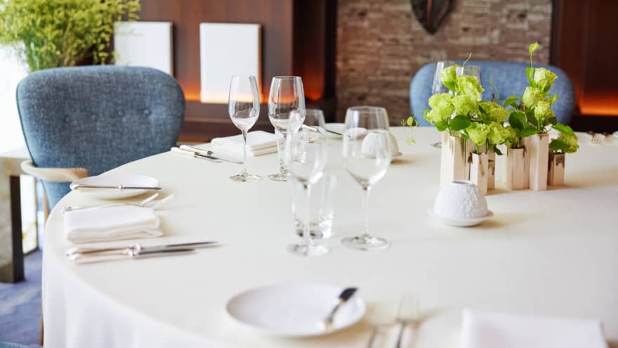Against a warm backdrop, round table set with white tablecloth, sparkling silver and wine glasses, fresh flowers at centre.