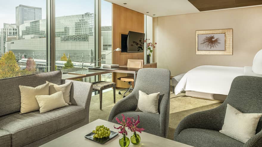 Junior suite with gray sofa, armchairs, work desk by a window with plaza view and king bed in the background